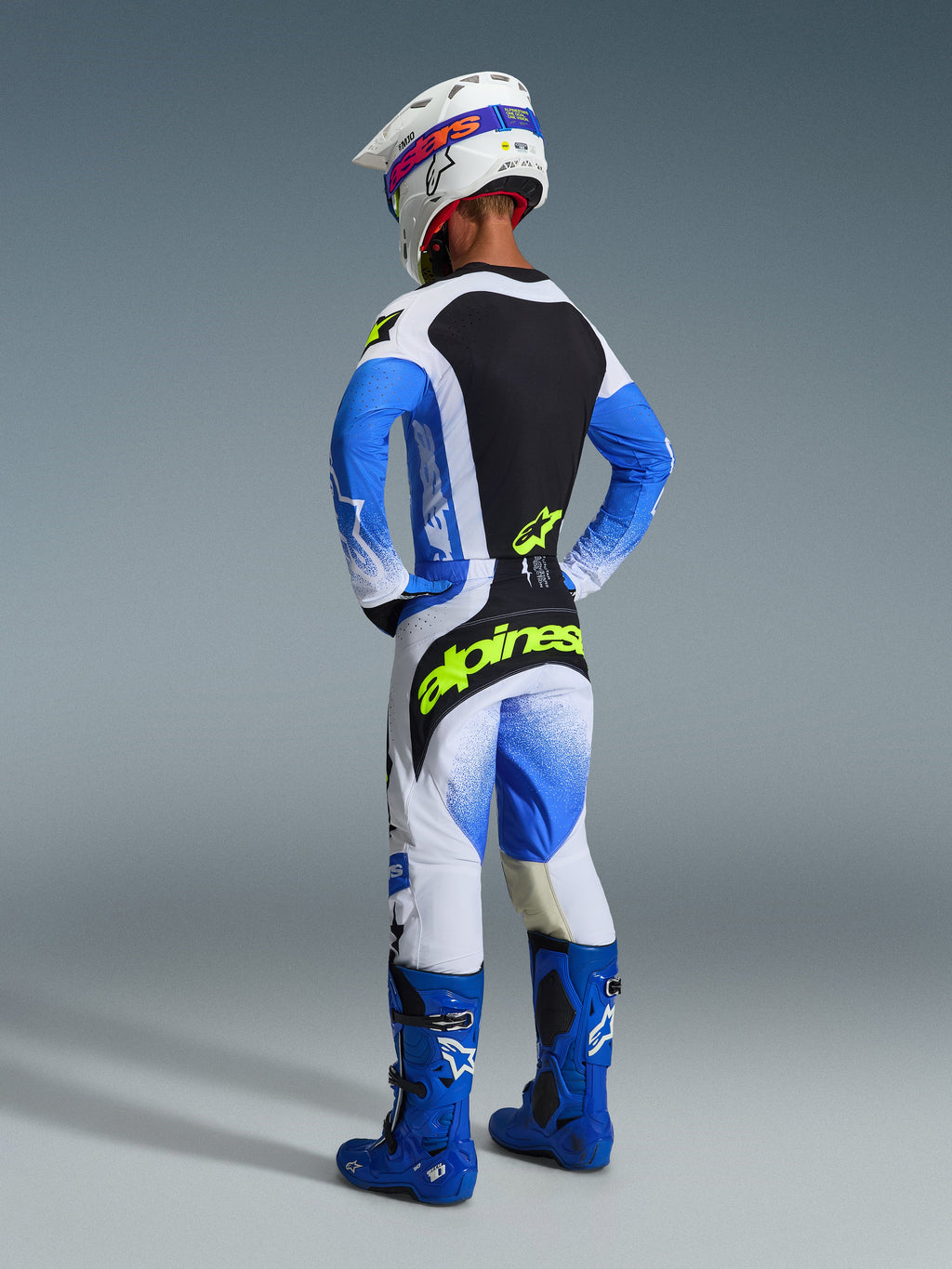 Motocross rider viewed from behind wearing the complete Alpinestars 2026 Techstar Knif Gear Set in Ucla Blue and Black, featuring a jersey with a black back panel and yellow fluo logo, matching blue and white pants with bold yellow branding, blue motocross boots, and a white racing helmet.