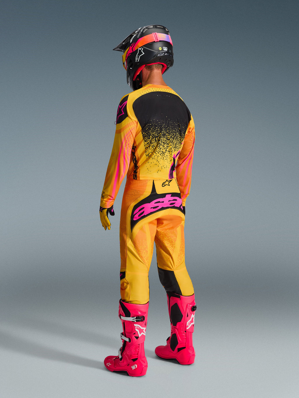 Motocross-Fahrer in der Rückansicht im Alpinestars 2026 Techstar Nomur Trikot, Orange, Gelb und Pink, passende Rennhose mit pinkem Branding, leuchtend pinke Stiefel und ein abgestimmter schwarz-pinker Helm, komplette Offroad-Wettkampfausrüstung