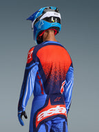 Motocross-Fahrer von hinten im 2026 Techstar Nomur Trikot, Dunkelblau mit orangefarbenen und UCLA-blauen Akzenten, mit einer großen strukturierten orangefarbenen Grafik auf dem Rücken, kombiniert mit einer passenden Motocross-Hose und einem blauen Rennhelm, professionell abgestimmte Ausrüstung für den Offroad-Sport.