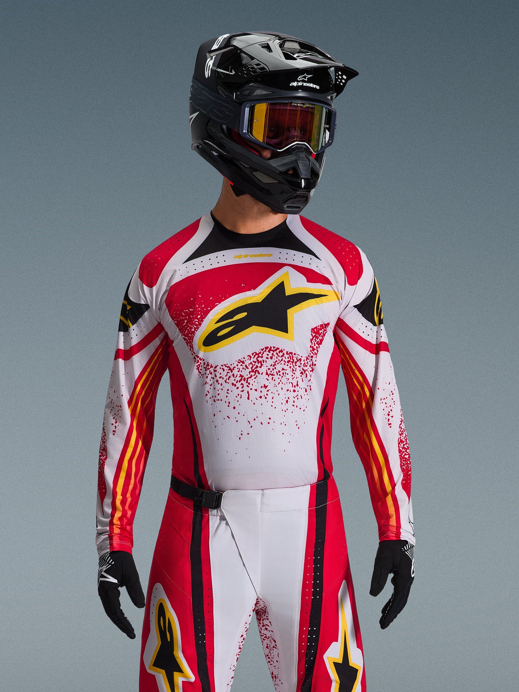 Alpinestars 2026 Techstar Nomur Trikot, Motocross-Trikot, Rot, Weiß und Schwarz, mit einem großen schwarzen A-Star-Logo auf der Brust und gelben Akzenten, rotes Splatter-Design, getragen mit einem schwarzen Helm und passender Offroad-Ausrüstung