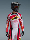 Alpinestars 2026 Techstar Nomur Trikot, Motocross-Trikot, Rot, Weiß und Schwarz, mit einem großen schwarzen A-Star-Logo auf der Brust und gelben Akzenten, rotes Splatter-Design, getragen mit einem schwarzen Helm und passender Offroad-Ausrüstung