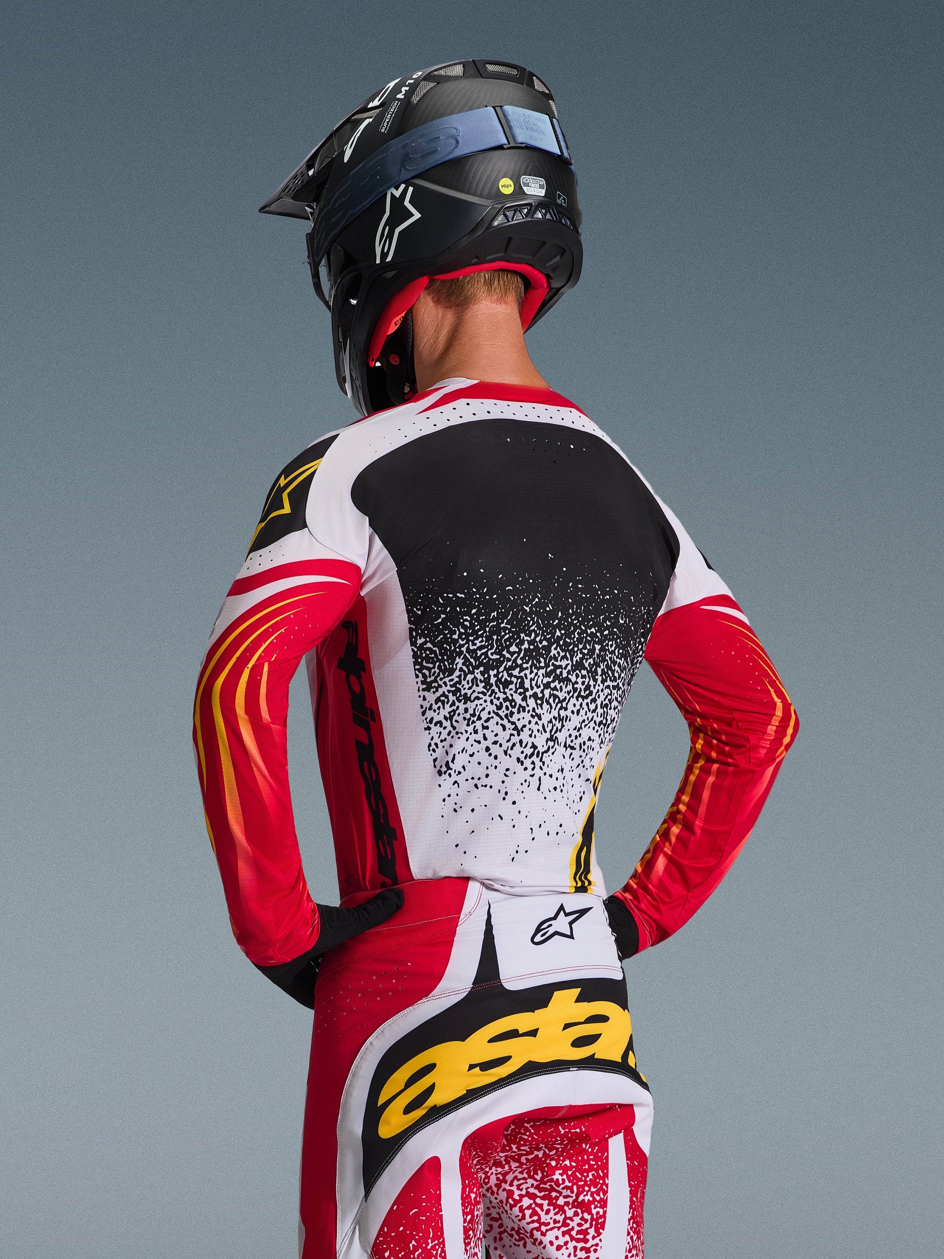 Motocross-Fahrer von hinten im Alpinestars 2026 Techstar Nomur Trikot und passender Hose, Rot, Weiß und Schwarz mit gelben Akzenten, gesprenkeltes Farbverlauf-Design auf dem Rücken, schwarzer Alpinestars-Helm und schwarze Handschuhe, professionelle Offroad-Rennausrüstung