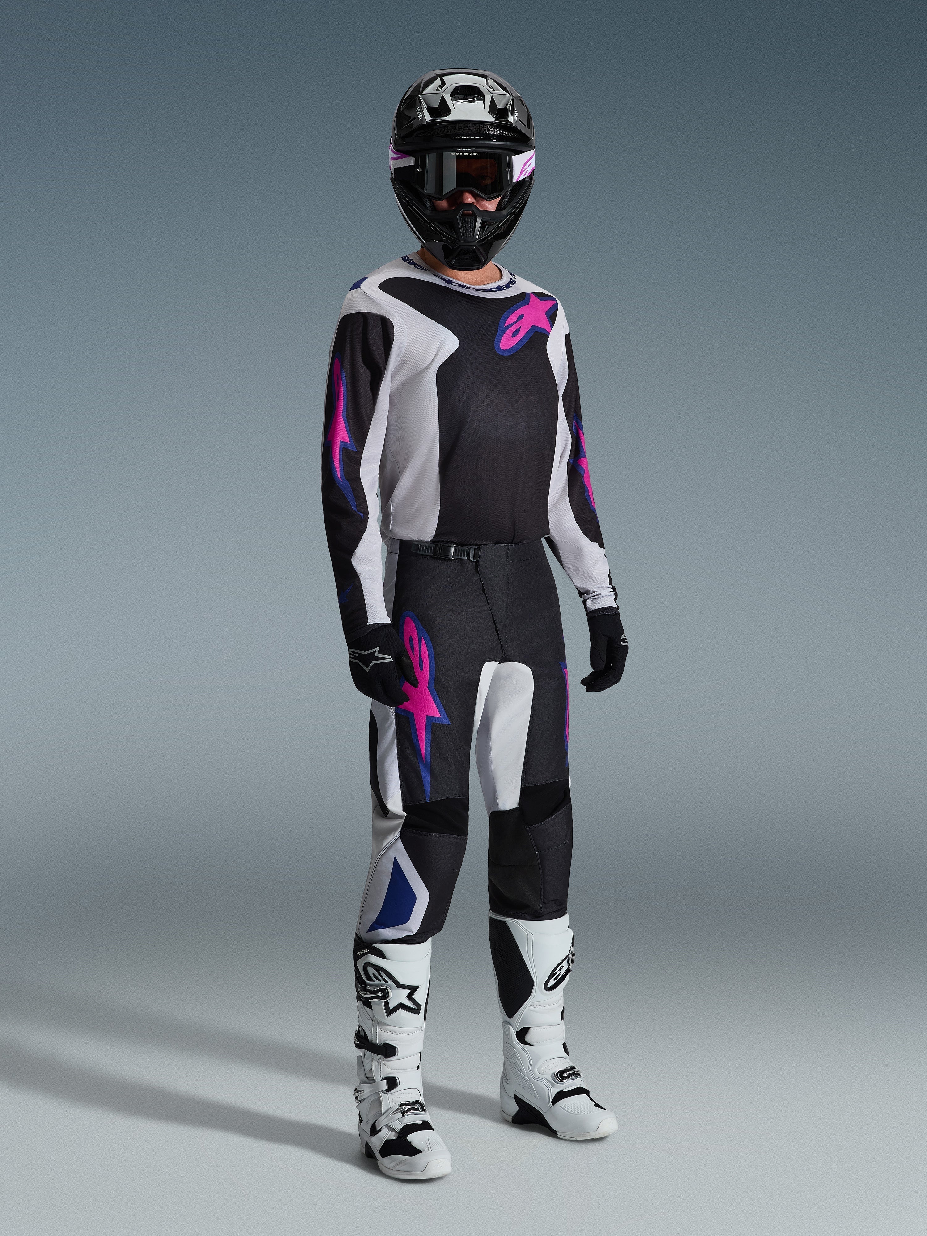 Motocross rider wearing complete racing gear, 2026 Fluid Grid Jersey in black with light gray and purple accents, matching Fluid Grid Pants, black helmet with coordinated goggles, white and black boots, and black gloves, professional studio setup for off-road racing.