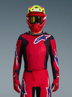 Alpinestars 2026 Fluid Grid Trikot, Motocross-Trikot, Rot, Schwarz und Lila, getragen von einem Fahrer mit einem roten und neongelben Helm und einer goldenen Goggle, mit großen Stern-Logos auf der Brust und den Ärmeln, abgestimmte Ausrüstung für den Offroad-Rennsport