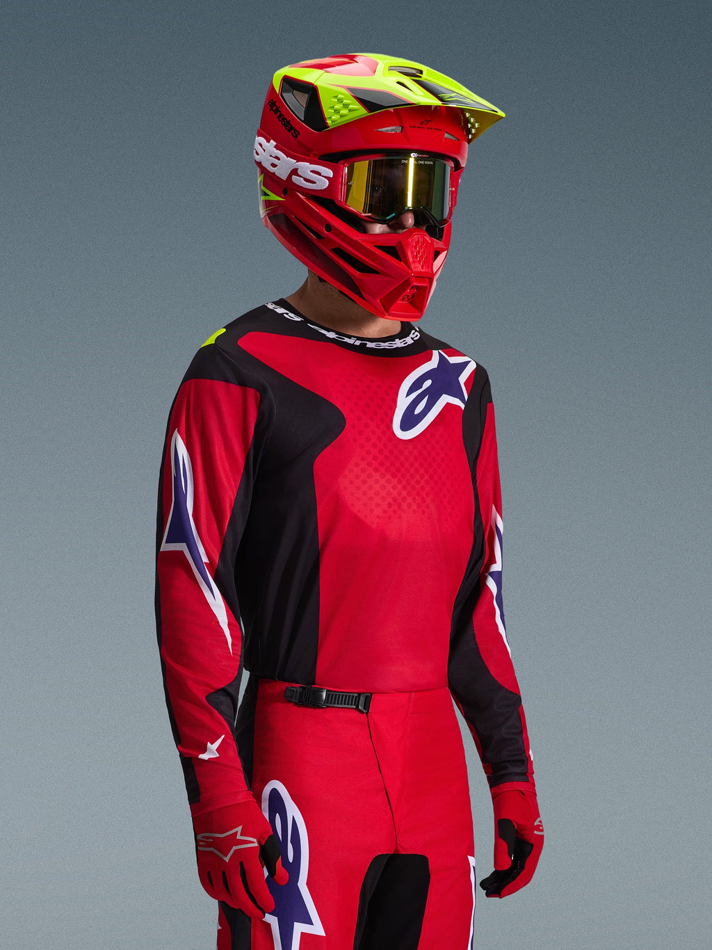 Motocross-Fahrer im Alpinestars 2026 Fluid Grid Trikot in Rot, Schwarz und Lila, kombiniert mit passender Motocross-Hose, rotem Helm mit neongelben Akzenten, goldener Goggle und roten Handschuhen, komplette Offroad-Rennausrüstung.