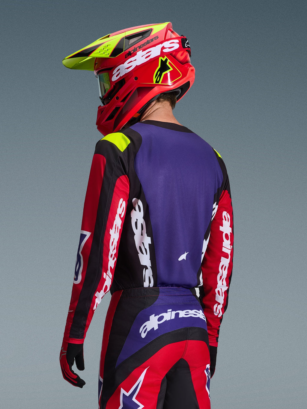 Motocross-Fahrer im Alpinestars 2026 Fluid Grid Trikot, Rot, Schwarz und Lila, Rückansicht mit lila Rückenpartie und roten Ärmeln mit weißem Branding, passende Hose und ein rot-neongelber Helm, koordiniertes Outfit für den Offroad-Rennsport