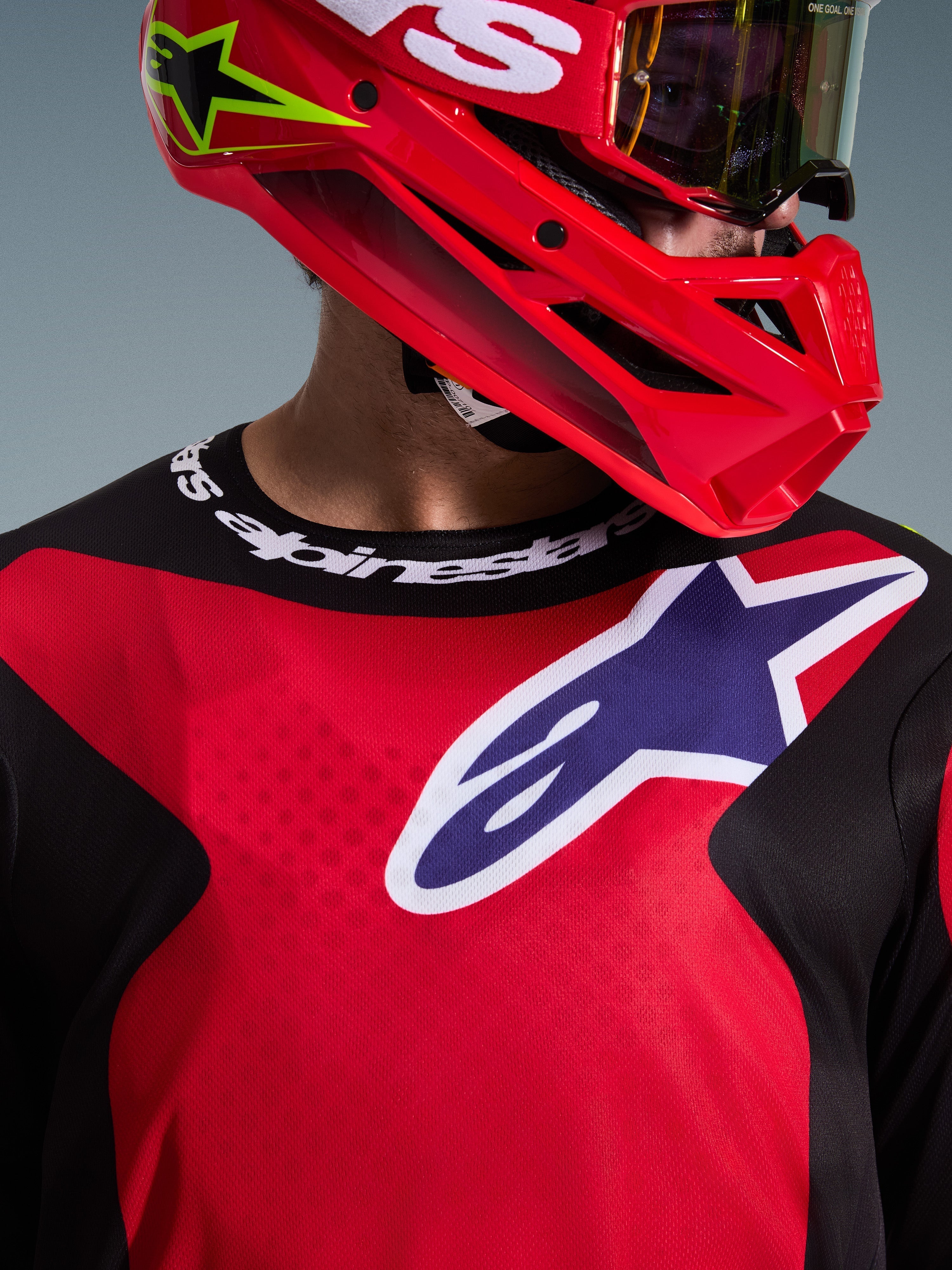 Nahaufnahme eines Motocross-Fahrers, der das 2026 Fluid Grid Trikot in Rot, Schwarz und Lila trägt, mit einem großen Alpinestars-Logo auf der Brust und einem Sportkragen mit Markenschriftzug, kombiniert mit einem roten Helm und einer Schutzbrille, was das Performance-Polygewebe und die sportliche Passform hervorhebt.