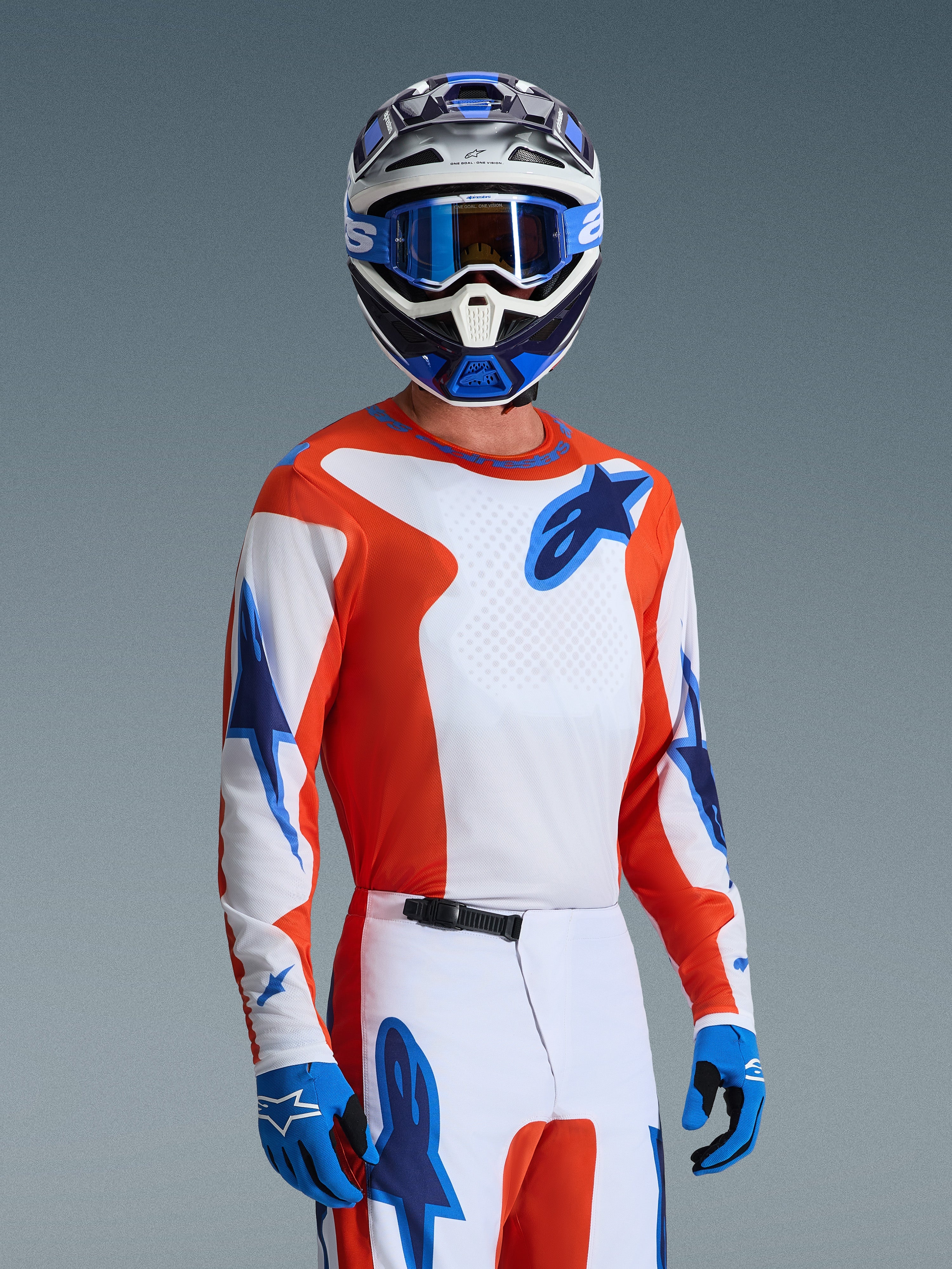 Alpinestars 2026 Fluid Grid Trikot, Motocross-Trikot in Orange UCLA Blue, getragen von einem Fahrer mit passender weiß-oranger Hose, blau-weißem Helm, blauer Brille und blauen Handschuhen, komplettes Offroad-Rennausrüstungs-Set