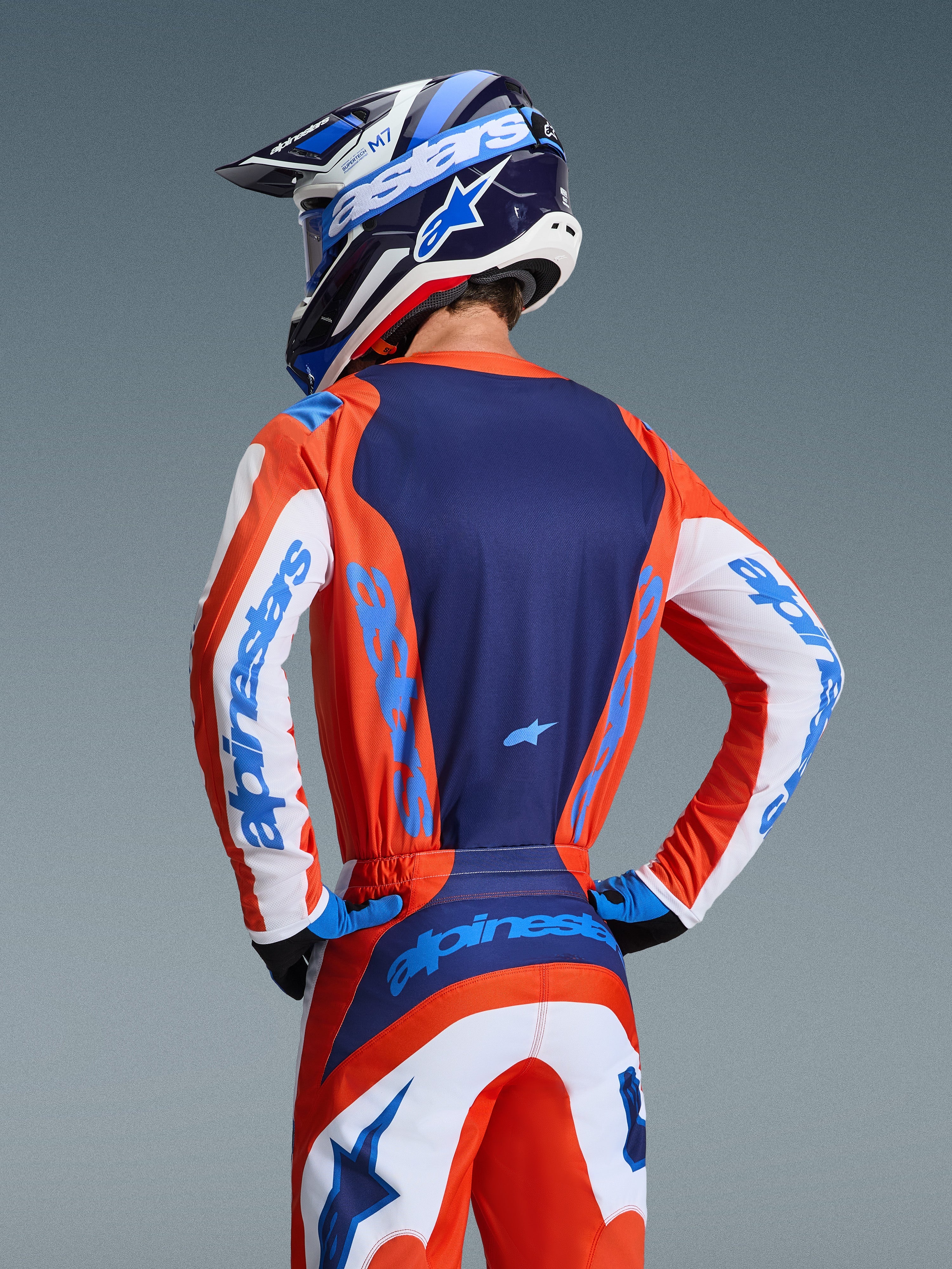 Alpinestars 2026 Fluid Grid Trikot, Rückansicht eines Motocross-Fahrers in abgestimmter Ausrüstung in Orange und UCLA-Blau, dunkelblaues Rückenteil, weiße Ärmelakzente mit blauem Branding, passender Helm, Performance-Offroad-Bekleidung