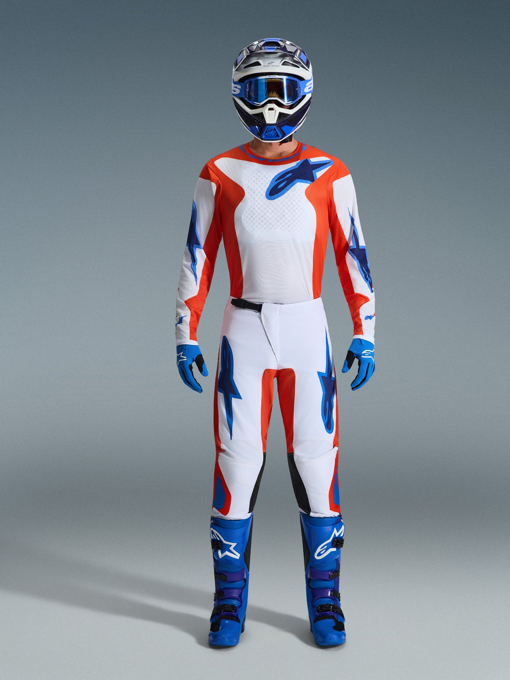 Motocross-Fahrer in kompletter Rennausrüstung, 2026 Fluid Grid Trikot in Orange und UCLA-Blau, passende weiße Hose mit orangefarbenen und blauen Akzenten, blau-weißer Helm mit blauer Schutzbrille, blau-violette Offroad-Stiefel, blaue Handschuhe, abgestimmtes Outfit für Motocross-Rennen