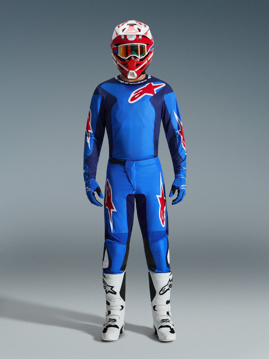 Motocross-Fahrer im Alpinestars 2026 Fluid Grid Trikot in Blau und Rot, passende blaue Hose mit weiß-roten Stern-Logos, rot-weißer Helm mit verspiegelter Schutzbrille, blaue Handschuhe und weiß-schwarze Rennstiefel, komplette Offroad-Ausrüstung.