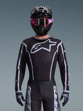 Alpinestars 2026 Fluid Apex Trikot, Motocross-Trikot, Schwarz und Grau, mit einem großen weißen Astars-Logo auf der Brust und grauen grafischen Akzenten an den Ärmeln, getragen von einem Fahrer mit pinker Brille und schwarzem Helm, koordinierte Offroad-Rennausrüstung