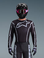 Alpinestars 2026 Fluid Apex Trikot, Motocross-Trikot, Schwarz und Grau, mit einem großen weißen Astars-Logo auf der Brust und grauen grafischen Akzenten an den Ärmeln, getragen von einem Fahrer mit pinker Brille und schwarzem Helm, koordinierte Offroad-Rennausrüstung
