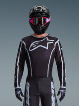 Alpinestars 2026 Fluid Apex Trikot, Motocross-Trikot, Schwarz und Grau, mit einem großen weißen Astars-Logo auf der Brust und grauen grafischen Akzenten an den Ärmeln, getragen von einem Fahrer mit pinker Brille und schwarzem Helm, koordinierte Offroad-Rennausrüstung