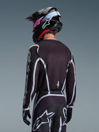 Motocross-Fahrer von hinten im Alpinestars 2026 Fluid Apex Trikot in Schwarz und Grau, passende Motocross-Hose mit weißen Akzenten, schwarzer Helm mit pinkem Branding, koordinierte Ausrüstung für Offroad-Rennsport