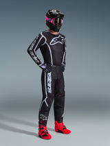 Motocross-Fahrer im 2026 Fluid Apex Trikot, Schwarz und Grau, kombiniert mit der passenden Hose, schwarzer Helm mit pinker Brille und leuchtend roten Stiefeln, markantes weißes Alpinestars-Branding auf der Brust und den Ärmeln, professionelles Offroad-Rennoutfit.