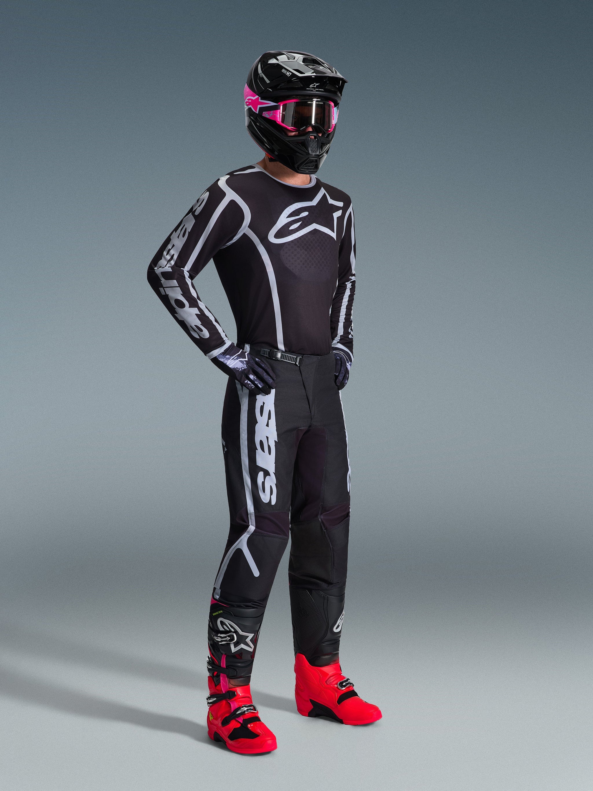 Motocross-Fahrer im 2026 Fluid Apex Trikot, Schwarz und Grau, kombiniert mit der passenden Hose, schwarzer Helm mit pinker Brille und leuchtend roten Stiefeln, markantes weißes Alpinestars-Branding auf der Brust und den Ärmeln, professionelles Offroad-Rennoutfit.