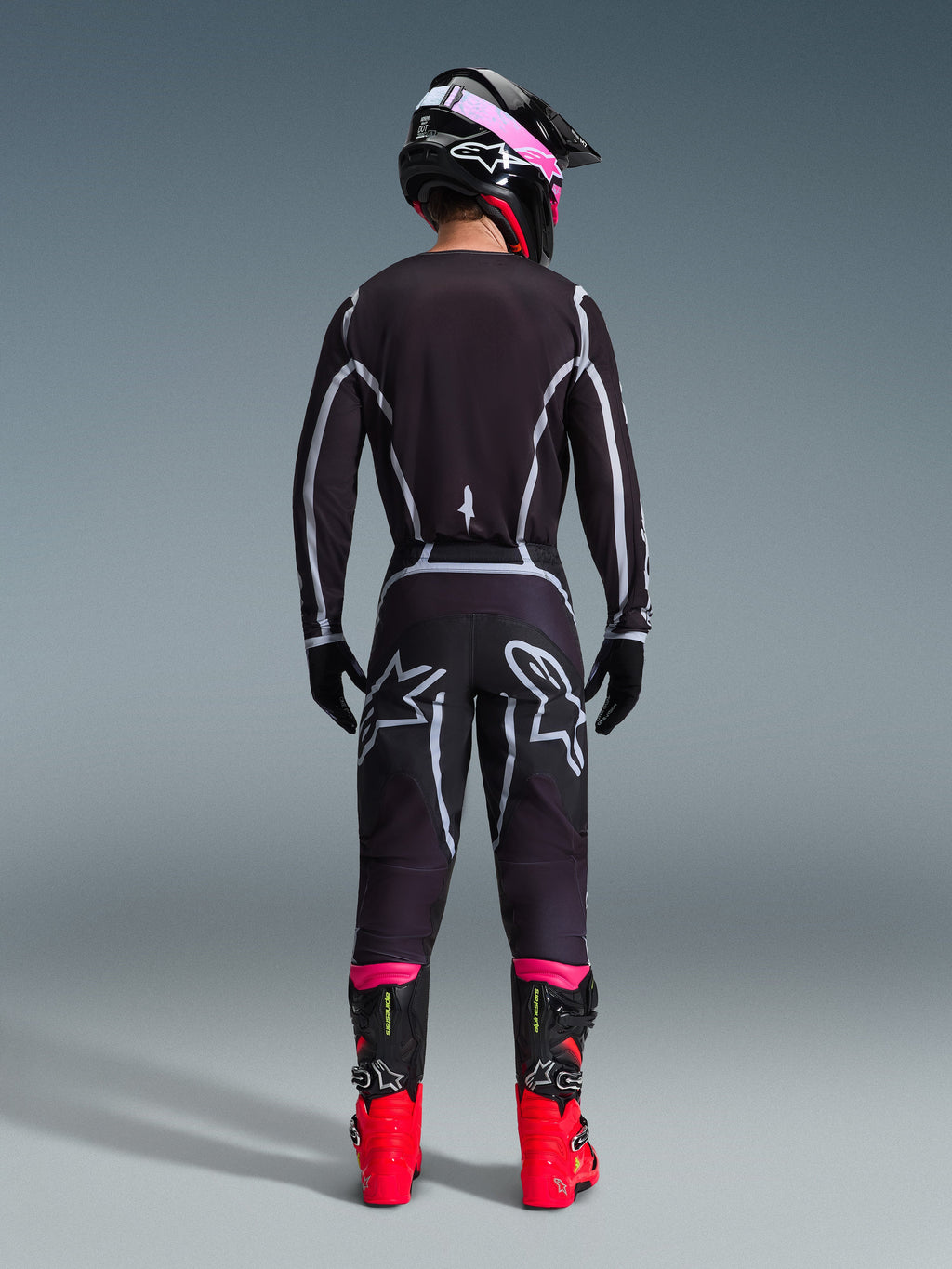 Motocross rider seen from behind wearing the Alpinestars 2026 Fluid Apex Gear Set, black jersey and pants with grey geometric lines and star logos, black helmet with pink accents, bright red and black boots, coordinated motocross racing gear