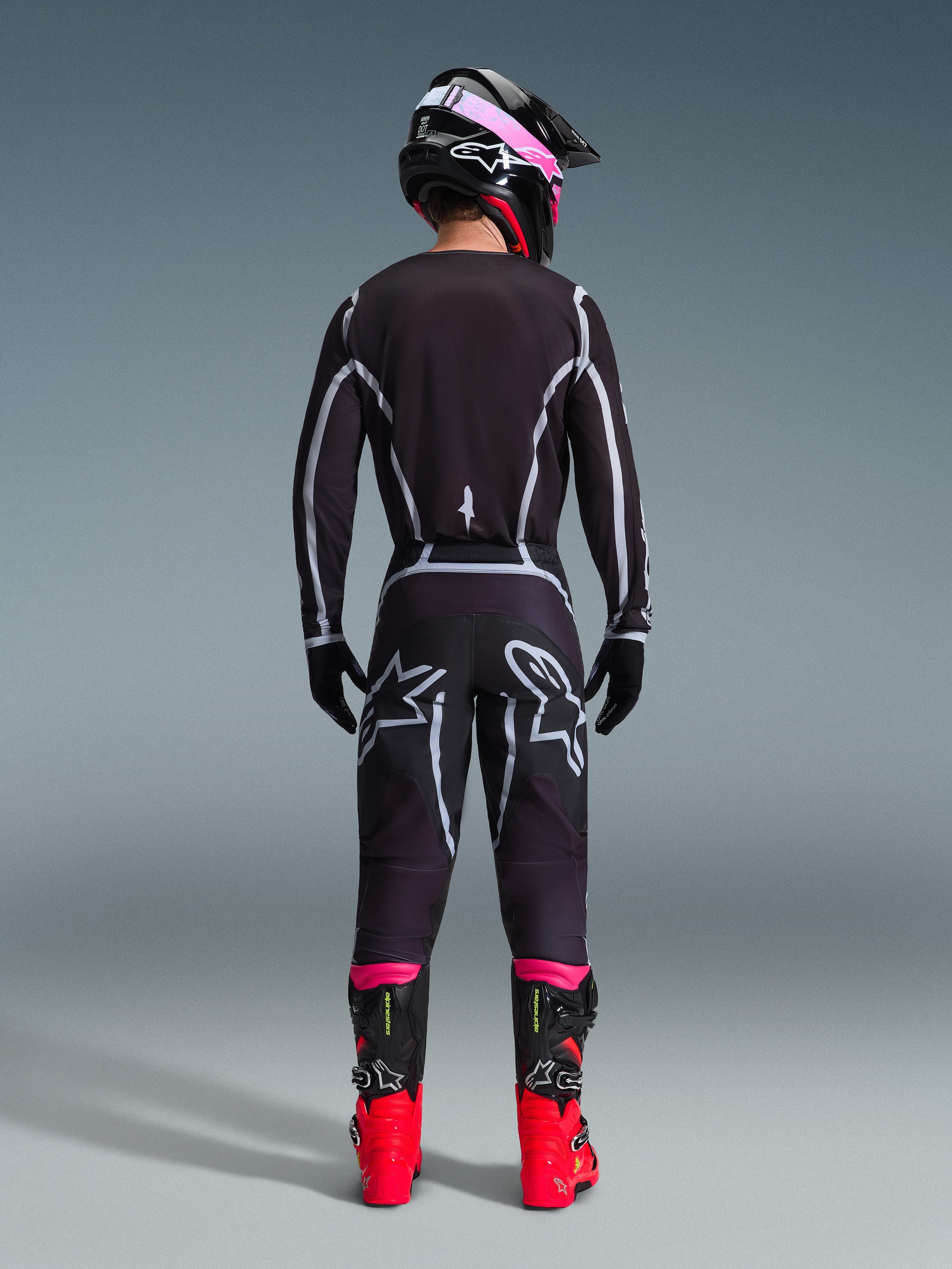 Motocross-Fahrer in Rückansicht, trägt das Alpinestars 2026 Fluid Apex Trikot und die passende Hose in Schwarz/Grau, mit weißen geometrischen Designlinien und Stern-Logos am Gesäß, kombiniert mit einem schwarz-pinken Helm und rot-schwarzen Rennstiefeln.