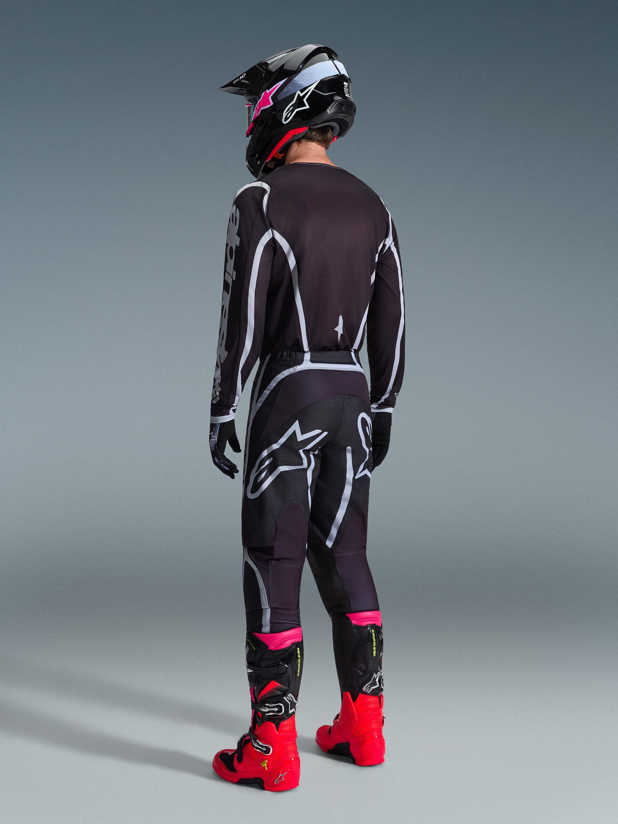 Motocross-Fahrer in kompletter Rennausrüstung, 2026 Fluid Apex Trikot in Schwarz und Grau von hinten gesehen, passende Hose mit weißen und grauen Akzenten, schwarzer Helm und leuchtend rote und pinkfarbene Stiefel, koordiniertes Outfit für Offroad-Motocross