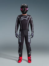 Motocross-Fahrer im Alpinestars 2026 Fluid Apex Trikot, Schwarz und Grau, großes weißes Astars-Logo auf der Brust, weiße geometrische Linien, passende schwarze Hose, schwarzer Helm mit pinker Brille und leuchtend pinke Stiefel, komplette Motocross-Rennausrüstung