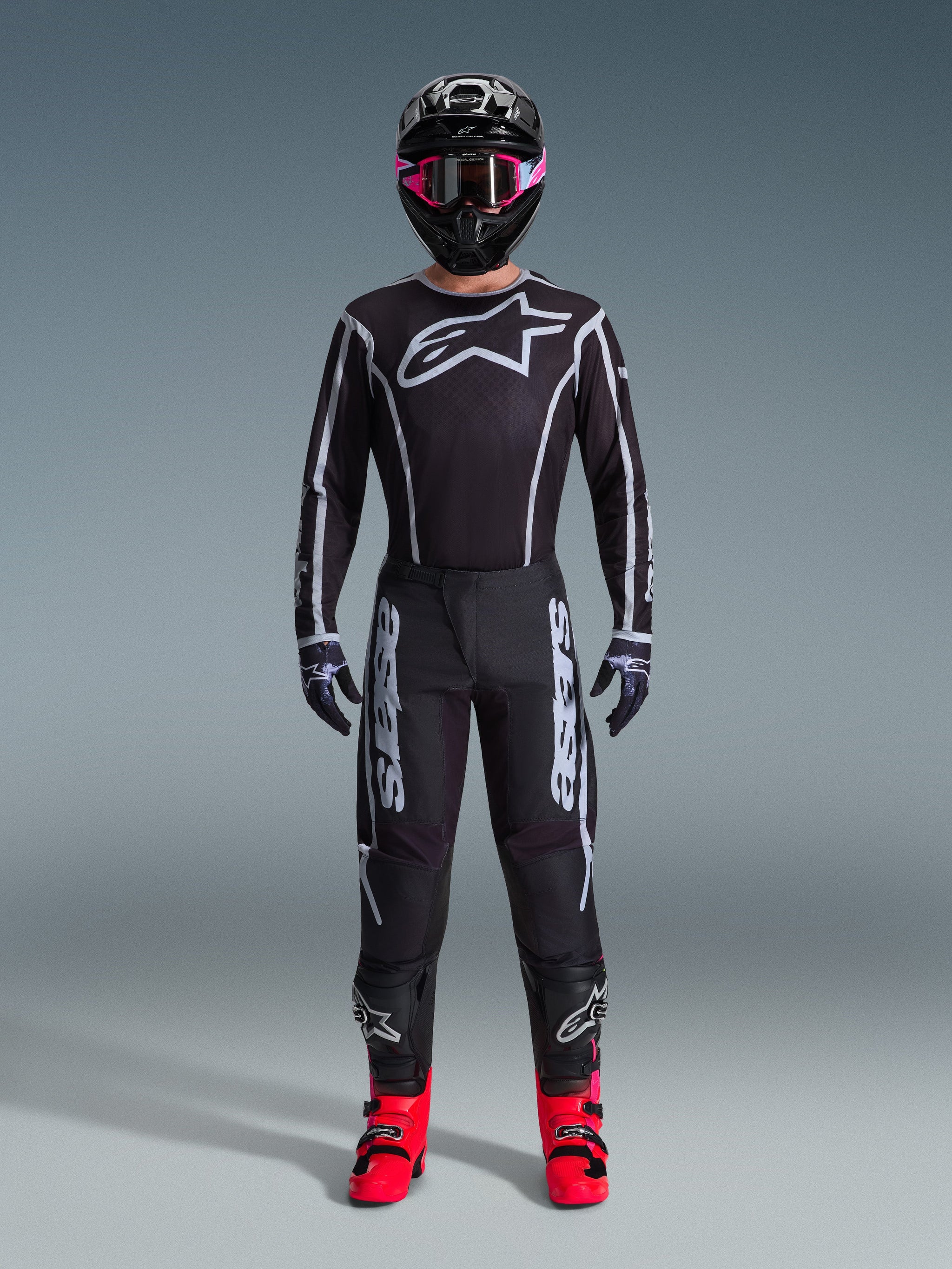 Motocross-Fahrer im Alpinestars 2026 Fluid Apex Trikot, Schwarz und Grau, großes weißes Astars-Logo auf der Brust, weiße geometrische Linien, passende schwarze Hose, schwarzer Helm mit pinker Brille und leuchtend pinke Stiefel, komplette Motocross-Rennausrüstung