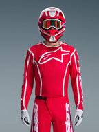 Alpinestars 2026 Fluid Apex Trikot, Motocross-Trikot, Rot und Weiß, getragen von einem Fahrer mit passendem rot-weißem Helm und orangefarbener Brille, mit markantem weißem Logo auf der Brust, sportliche Passform, Teil einer abgestimmten Offroad-Rennausrüstung
