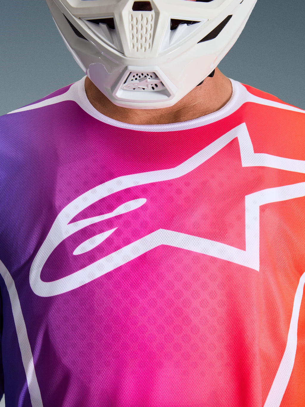 Alpinestars 2026 Fluid Apex Trikot, Motocross-Trikot, mehrfarbiger Farbverlauf in Lila, Pink und Orange, großes weißes Astars-Logo auf der Brust, Nahaufnahme getragen mit einem weißen Motocross-Helm