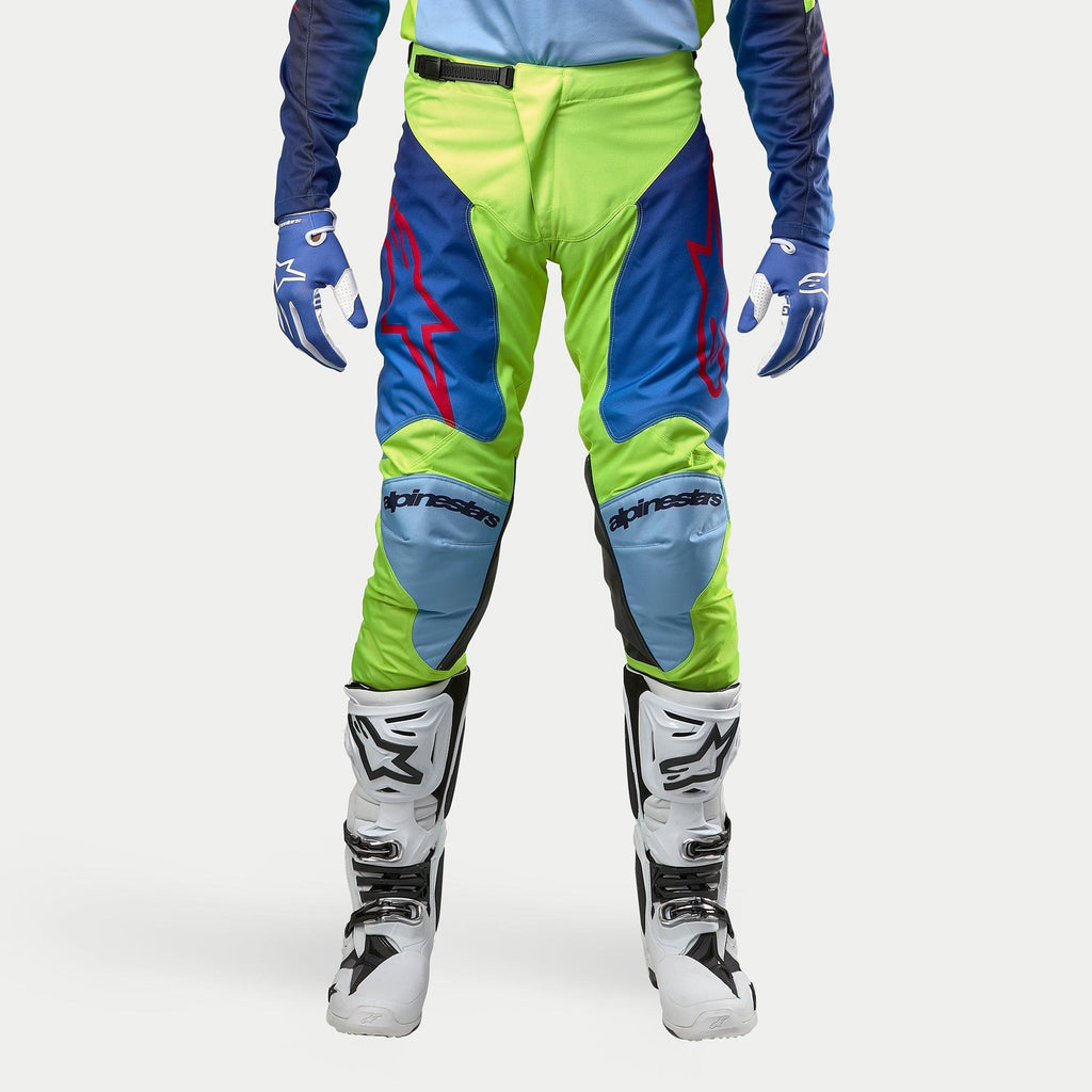 Alpinestars 2024 Racer Hoen Hose, Motocross-Hose, Gelb Fluo und Blau Night Navy, getragen mit weiß-schwarzen Motocross-Stiefeln und blauen Handschuhen, mit markanten roten Logos, hellblauen Knieeinsätzen und einem sportlichen ergonomischen Design für den Offroad-Einsatz.