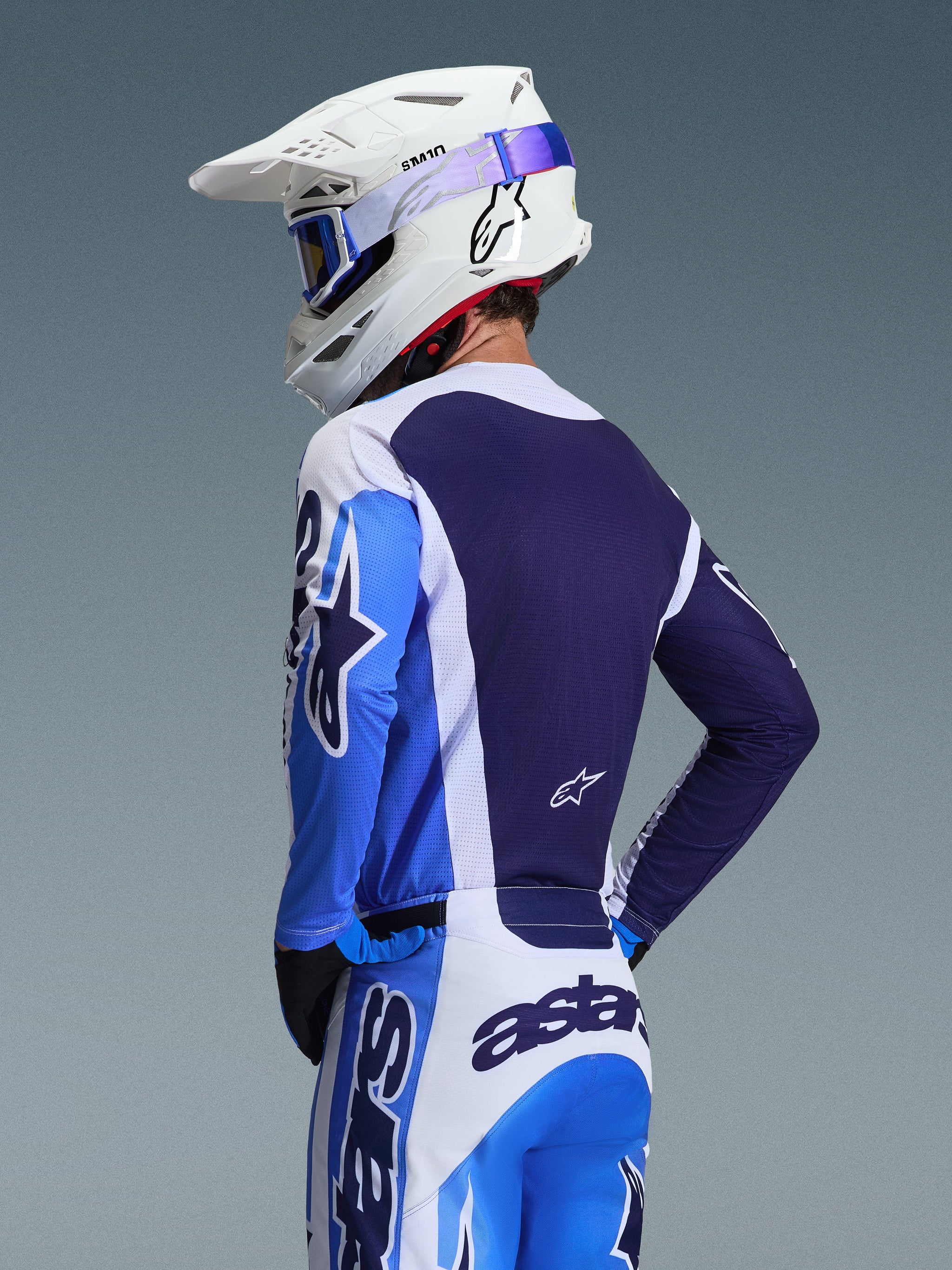 Motocross-Fahrer im Alpinestars 2026 Racer Air Portl Trikot in Ucla Blue und Weiß, Rückansicht mit Fokus auf das atmungsaktive marineblaue Mesh-Rückenteil, kombiniert mit passender Hose, weißem Alpinestars-Helm und blauer Brille, abgestimmtes Motocross-Ausrüstungsset.