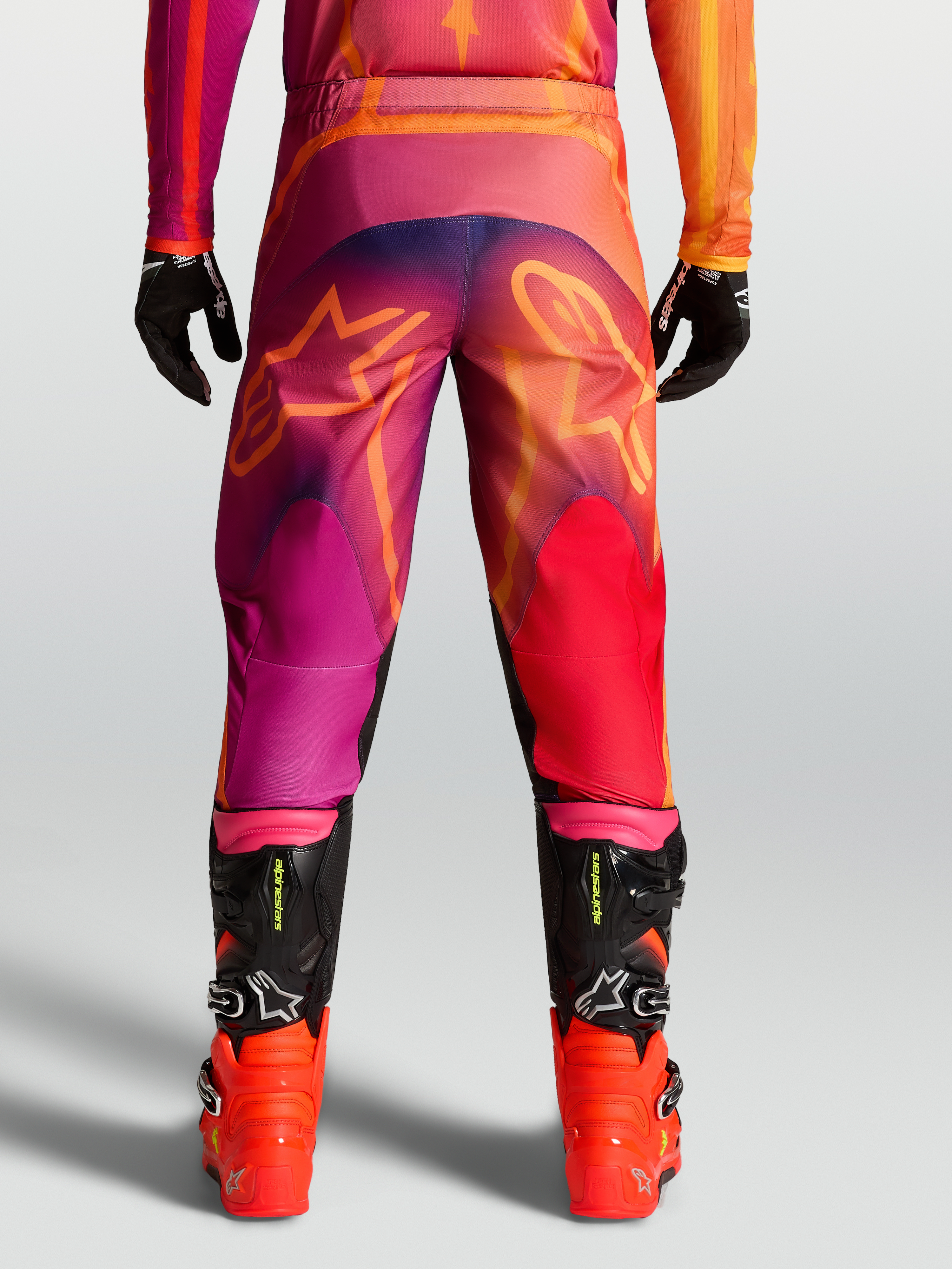 Alpinestars Fluid Kepler Hose, Motocross-Hose für Herren, Lila und Orange, mit Stern-Grafiken am Gesäß und schwarzen MX-Stiefeln.