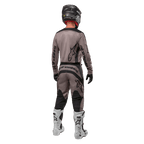 Alpinestars 2024 Fluid Lurv Trikot, Motocross-Trikot, Mud Black, Rückansicht eines Motocross-Fahrers in passender Rennausrüstung, schwarzer Helm und weiße Stiefel, markante schwarze Alpinestars-Logos, sportliche Passform für Offroad-Performance.