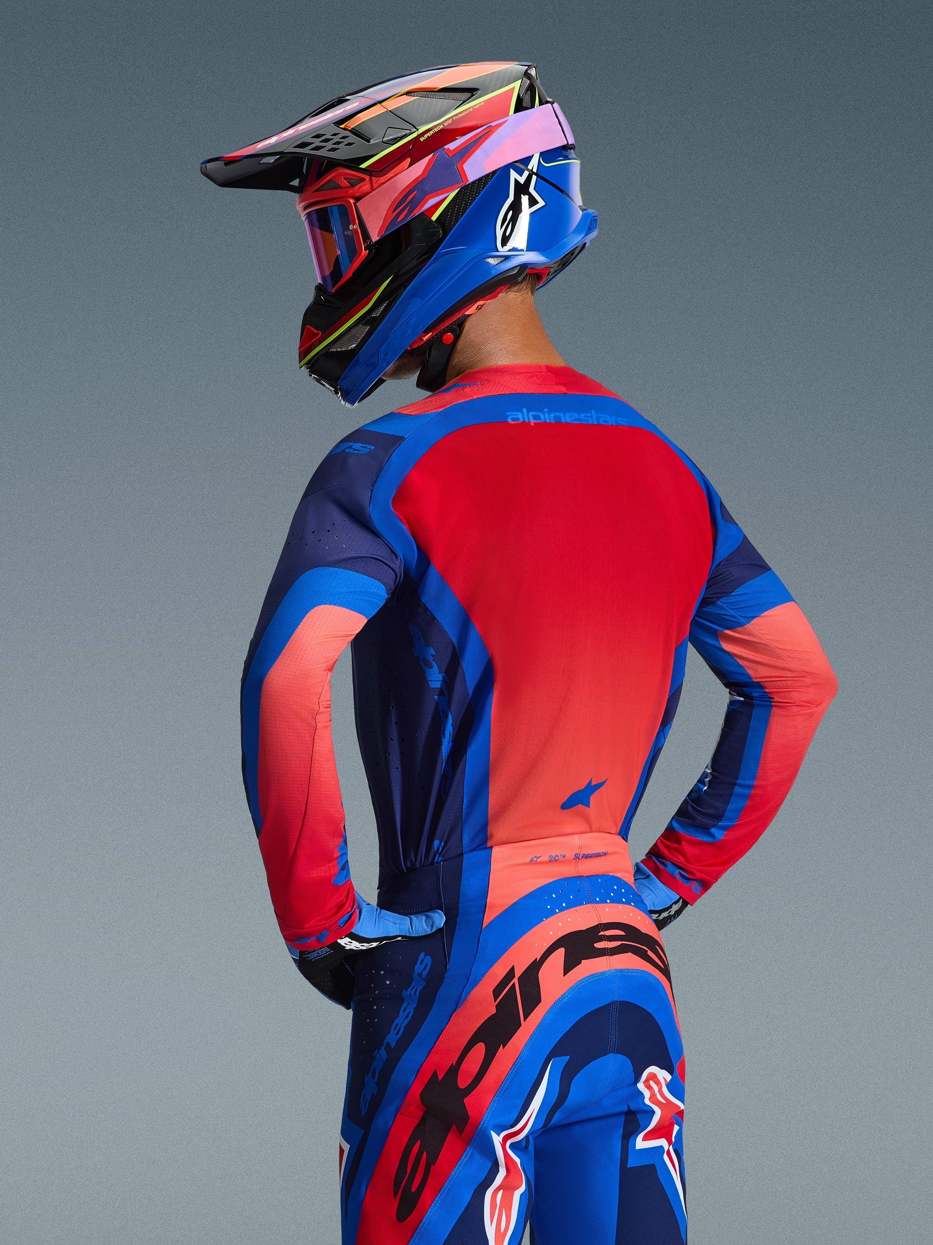 Rear view of a motocross rider wearing the Alpinestars 2026 Supertech Vista Jersey in Dark Navy, Hot Coral, and Cobalt, featuring a vibrant red back panel and seamless construction, paired with matching pants and helmet for a complete racing look.