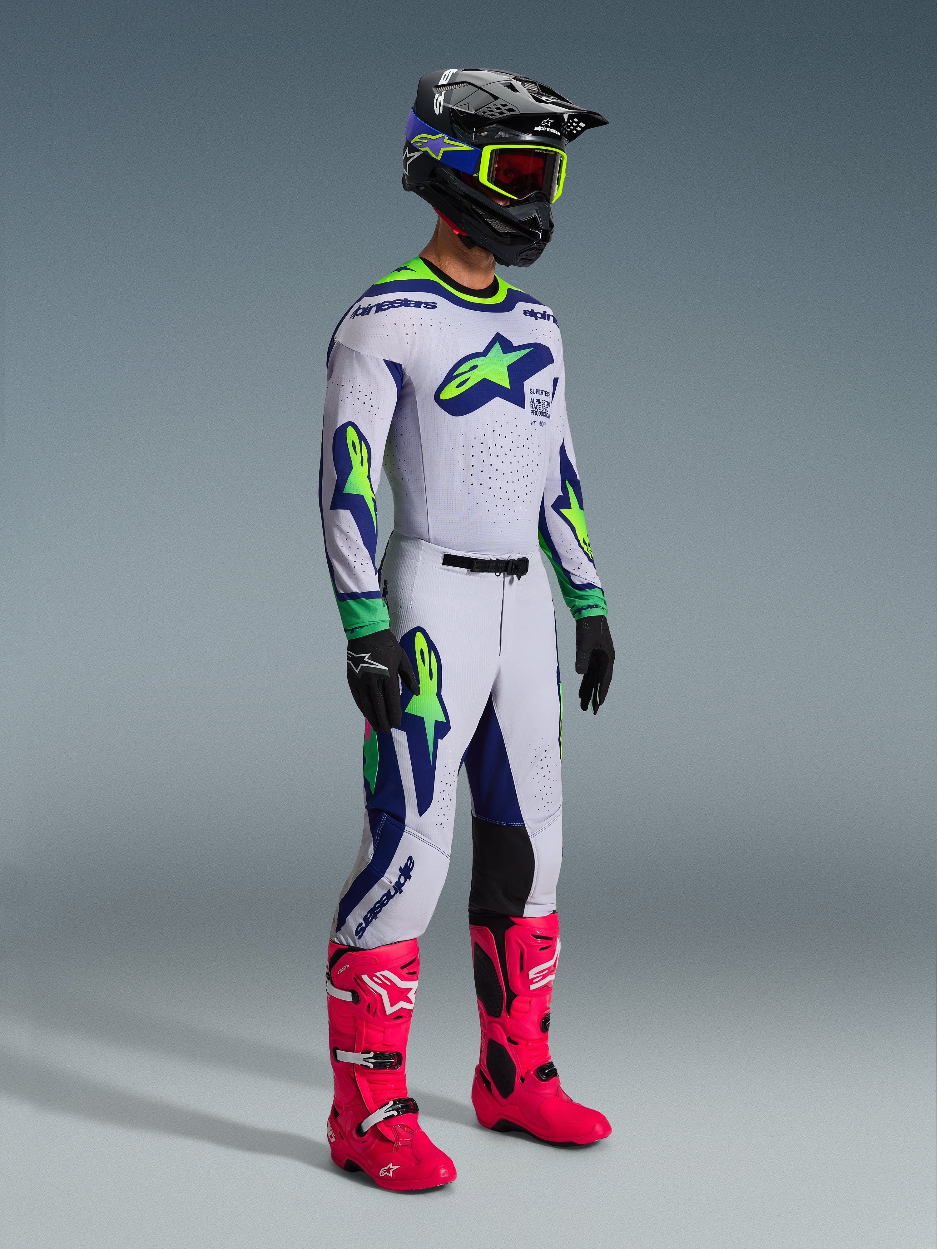 Motocross rider wearing the Alpinestars 2026 Supertech Vista Jersey in Light Gray Yellow Fluo Blue, matching racing pants, bright pink boots, and a black helmet with yellow goggles, featuring laser-cut ventilation and a professional athletic fit for off-road racing