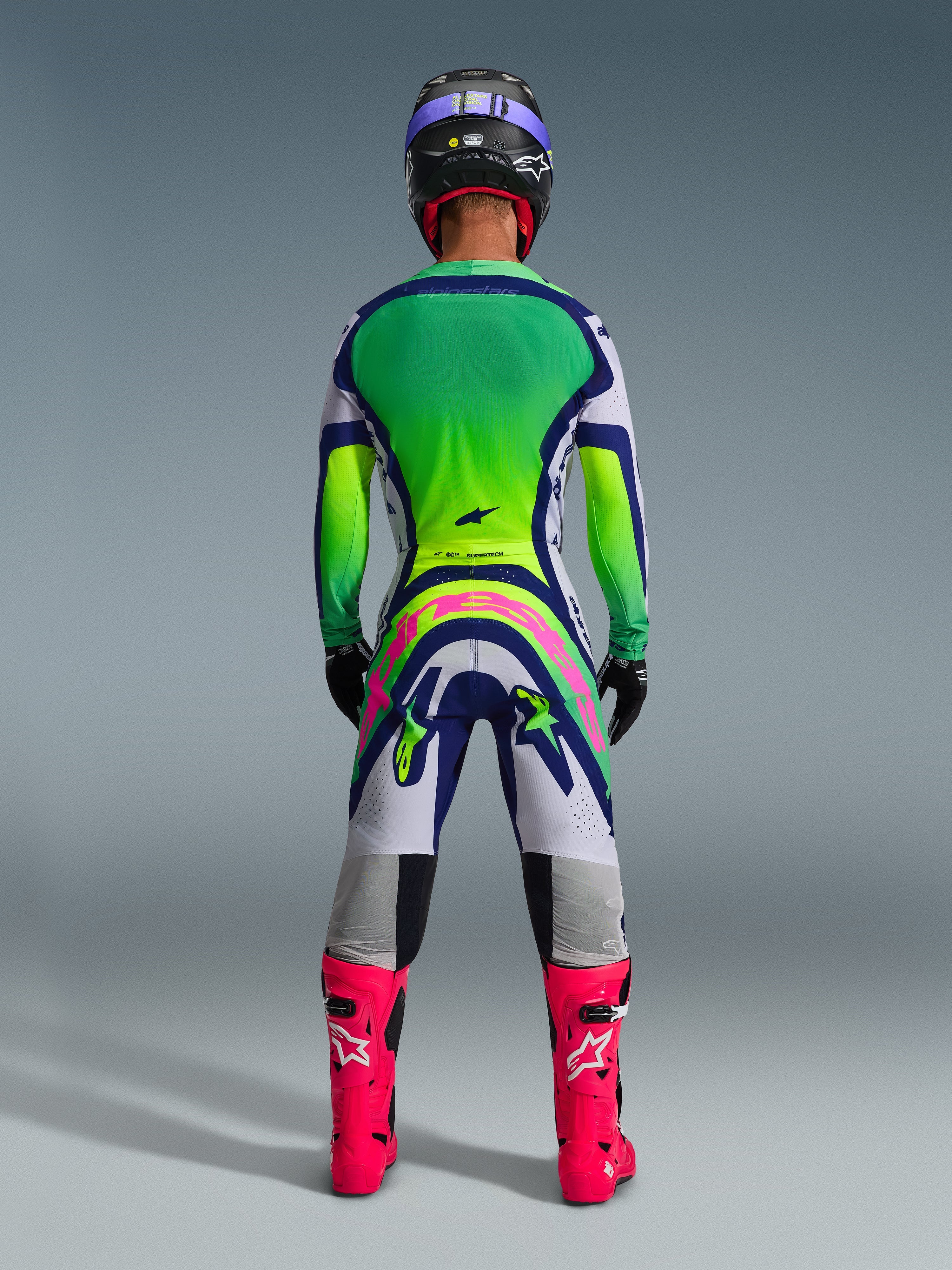 Motocross rider wearing complete racing gear from behind, featuring the 2026 Supertech Vista Jersey with a green and yellow gradient back, white and blue pants with large pink Alpinestars branding, bright pink boots, and a black helmet with purple accents, coordinated professional outfit for off-road racing.