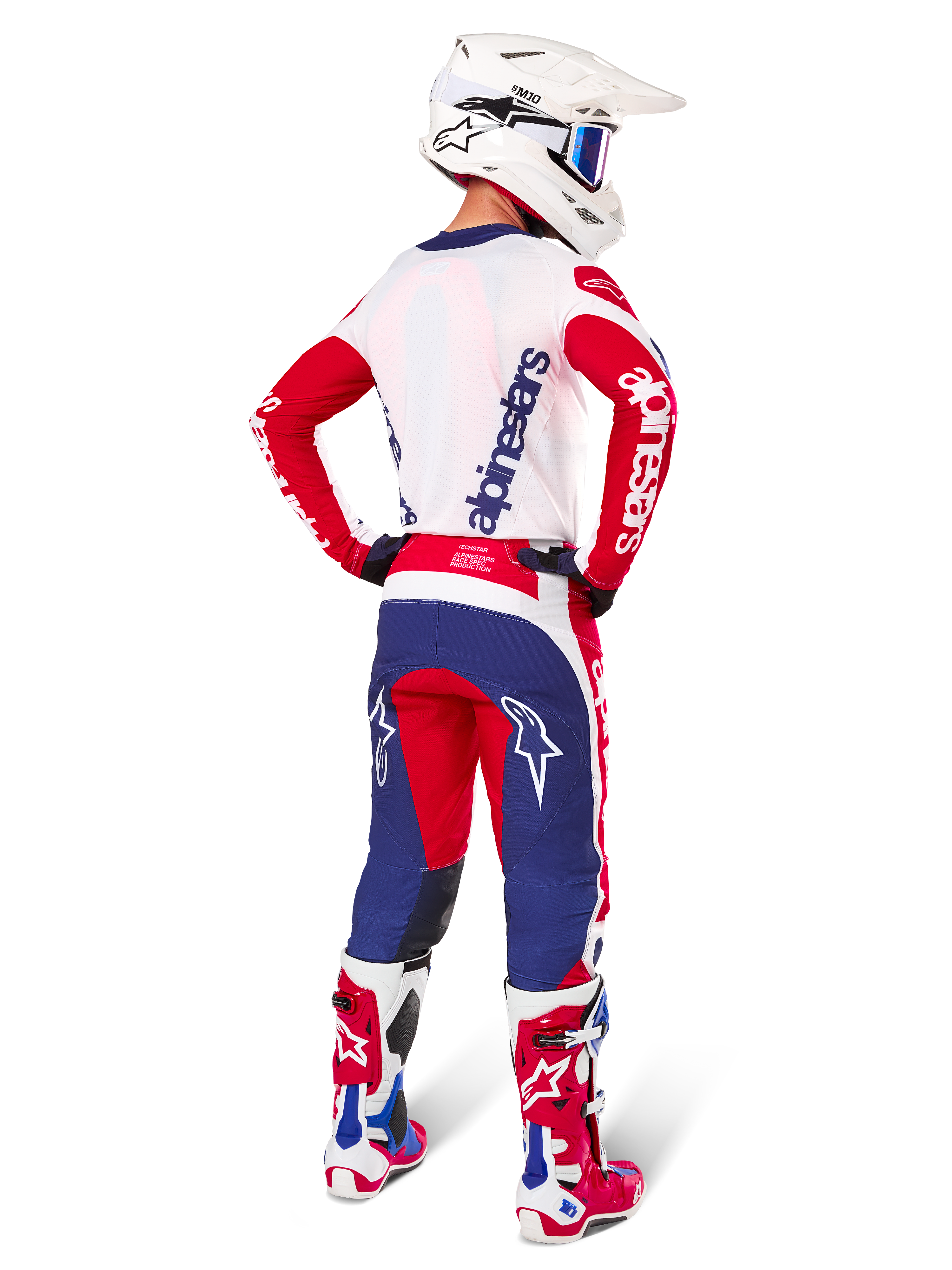 Motocross-Fahrer in der Rückansicht, trägt das Alpinestars Limited Edition Techstar Fortitude Trikot und Ausrüstungsset in der Farbkombination Bright Red White Blue, mit weißem Alpinestars-Branding auf den roten Ärmeln und einem marineblauen Logo auf dem weißen Rücken, kombiniert mit einem weißen Motocross-Helm und passenden Rennstiefeln.