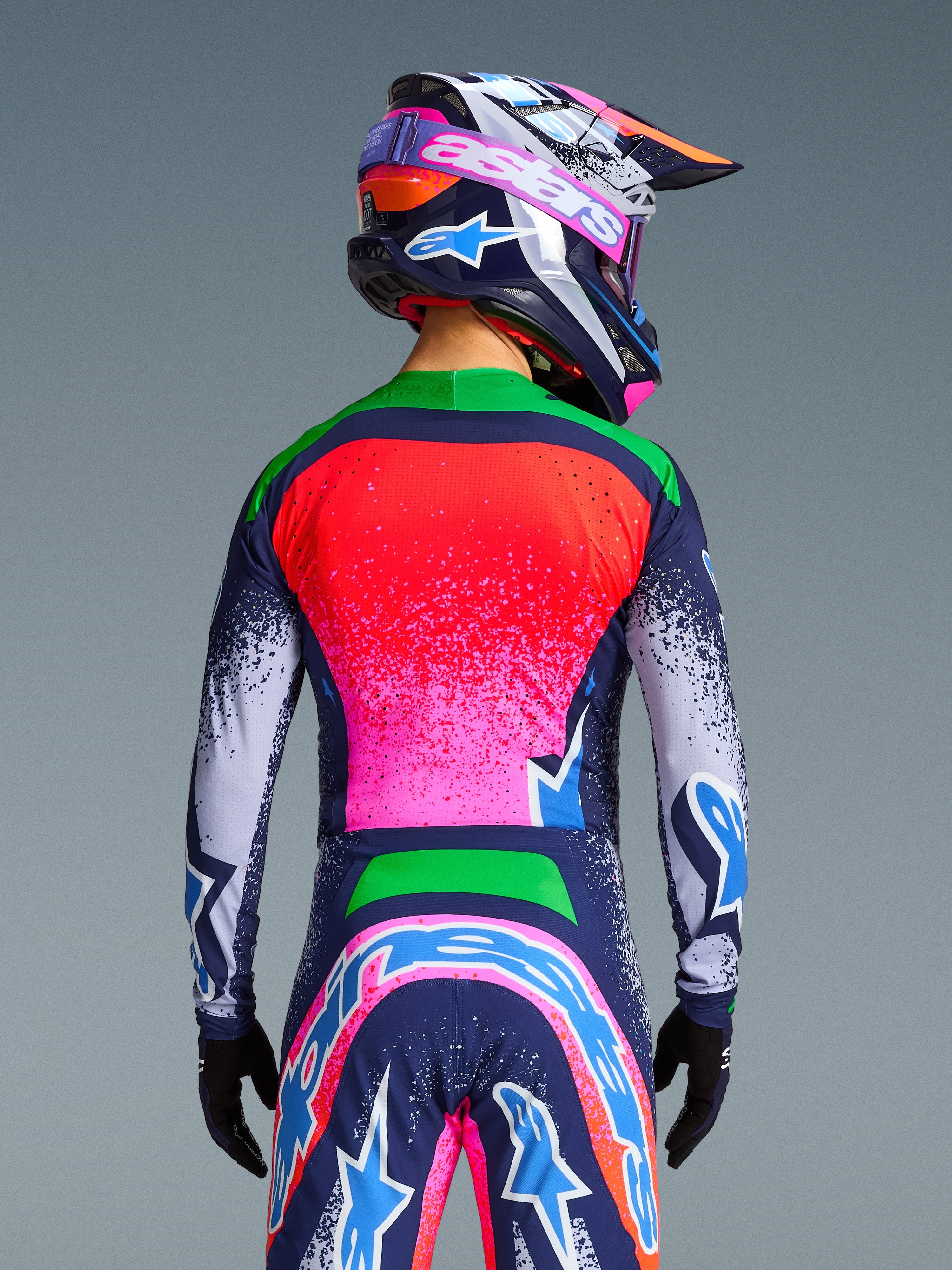 Alpinestars Limited Edition Supertech Records Trikot, Rückansicht eines Motocross-Fahrers in einer lebhaften, mehrfarbigen Ausrüstung, mit einem pink-roten Farbverlauf am Rücken, grünen Schultern und marineblau gesprenkelten Ärmeln, kombiniert mit einem passenden Helm und einer passenden Hose für einen professionellen Offroad-Rennlook.