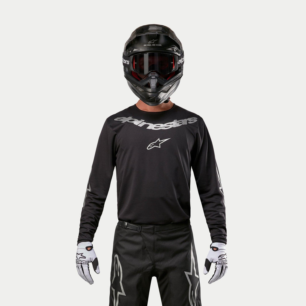 Alpinestars 2024 Fluid Graphite Jersey, schwarz-silbernes Motocross-Jersey, getragen von einem Fahrer mit schwarzem Helm und roter Brille, mit silbernem Alpinestars-Branding auf der Brust, kombiniert mit einer schwarzen Hose und weißen Handschuhen für Offroad-Performance.