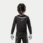 Alpinestars 2024 Fluid Graphite Jersey, schwarz-silbernes Motocross-Jersey, getragen von einem Fahrer mit schwarzem Helm und roter Brille, mit silbernem Alpinestars-Branding auf der Brust, kombiniert mit einer schwarzen Hose und weißen Handschuhen für Offroad-Performance.