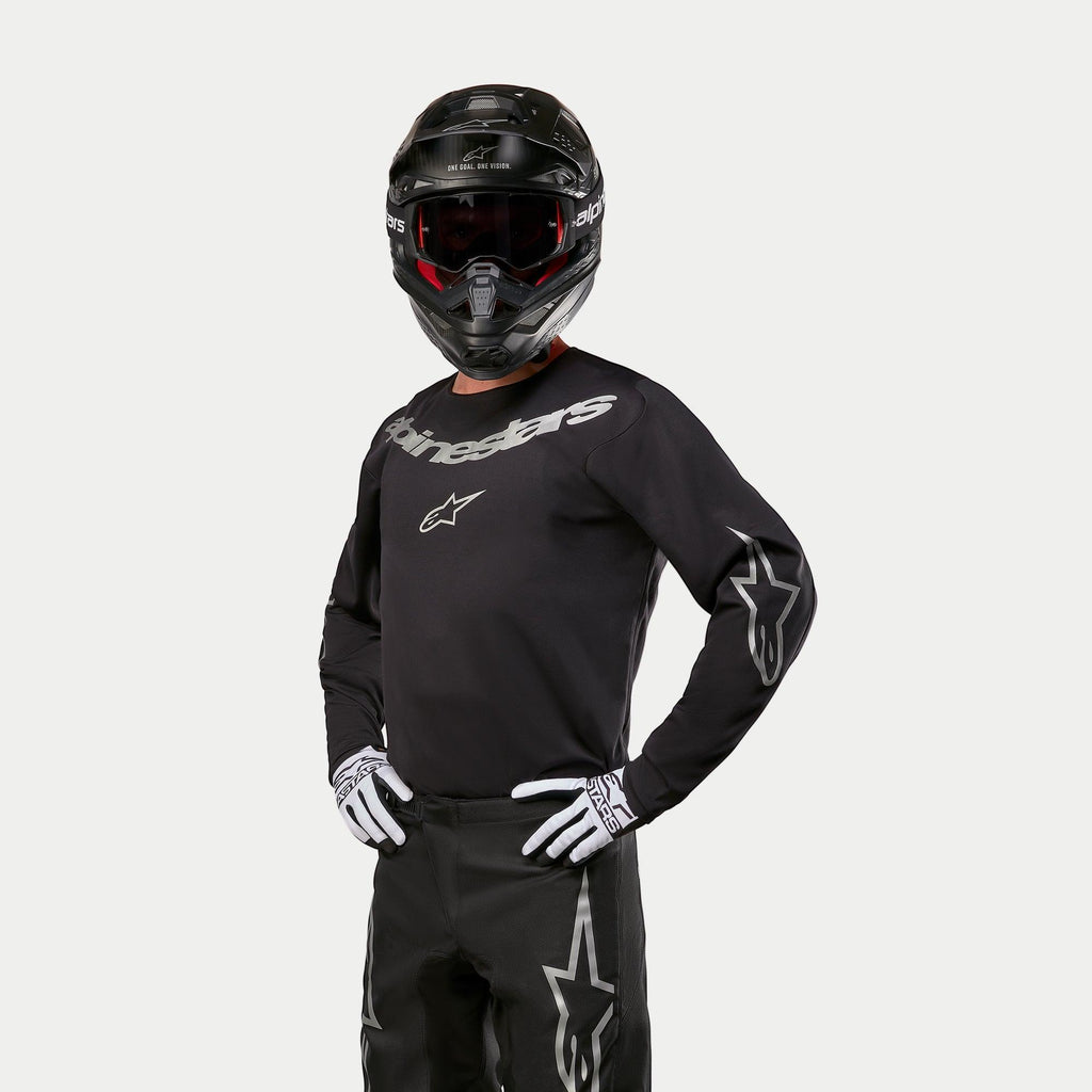 Motocross-Fahrer im Alpinestars 2024 Fluid Graphite Trikot, Schwarz und Silber, kombiniert mit einer schwarzen Motocross-Hose, einem schwarzen Helm mit rot getönter Brille und weiß-schwarzen Handschuhen, mit silbernen Markenlogos auf der Brust und den Ärmeln.