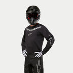 Motocross-Fahrer im Alpinestars 2024 Fluid Graphite Trikot, Schwarz und Silber, kombiniert mit einer schwarzen Motocross-Hose, einem schwarzen Helm mit rot getönter Brille und weiß-schwarzen Handschuhen, mit silbernen Markenlogos auf der Brust und den Ärmeln.