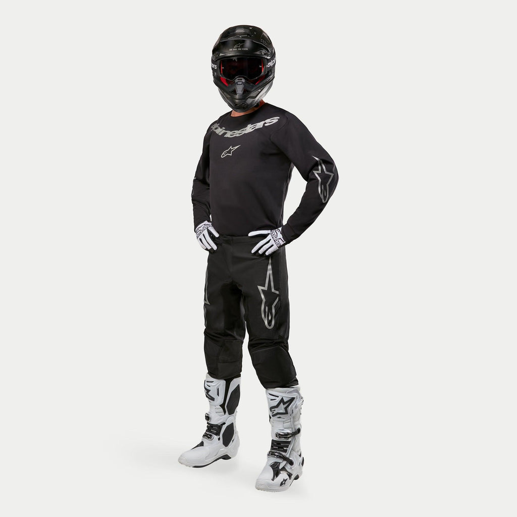Motocross-Fahrer mit Alpinestars 2024 Fluid Graphite Trikot in Schwarz und Silber, passender schwarzer Hose mit silbernen Stern-Logos, weiß-schwarzen Stiefeln, weiß-schwarzen Handschuhen und einem schwarzen Helm mit roter Brille, komplette Offroad-Rennausrüstung