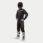 Motocross-Fahrer mit Alpinestars 2024 Fluid Graphite Trikot in Schwarz und Silber, passender schwarzer Hose mit silbernen Stern-Logos, weiß-schwarzen Stiefeln, weiß-schwarzen Handschuhen und einem schwarzen Helm mit roter Brille, komplette Offroad-Rennausrüstung