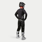Motocross-Fahrer im Alpinestars 2024 Fluid Graphite Trikot in Schwarz Silber aus der Rückansicht, langärmliges Jersey in athletischer Passform kombiniert mit schwarzer Motocross-Hose, schwarzem Helm und weiß-schwarzen Stiefeln, komplette professionelle Ausrüstung für den Offroad-Rennsport.