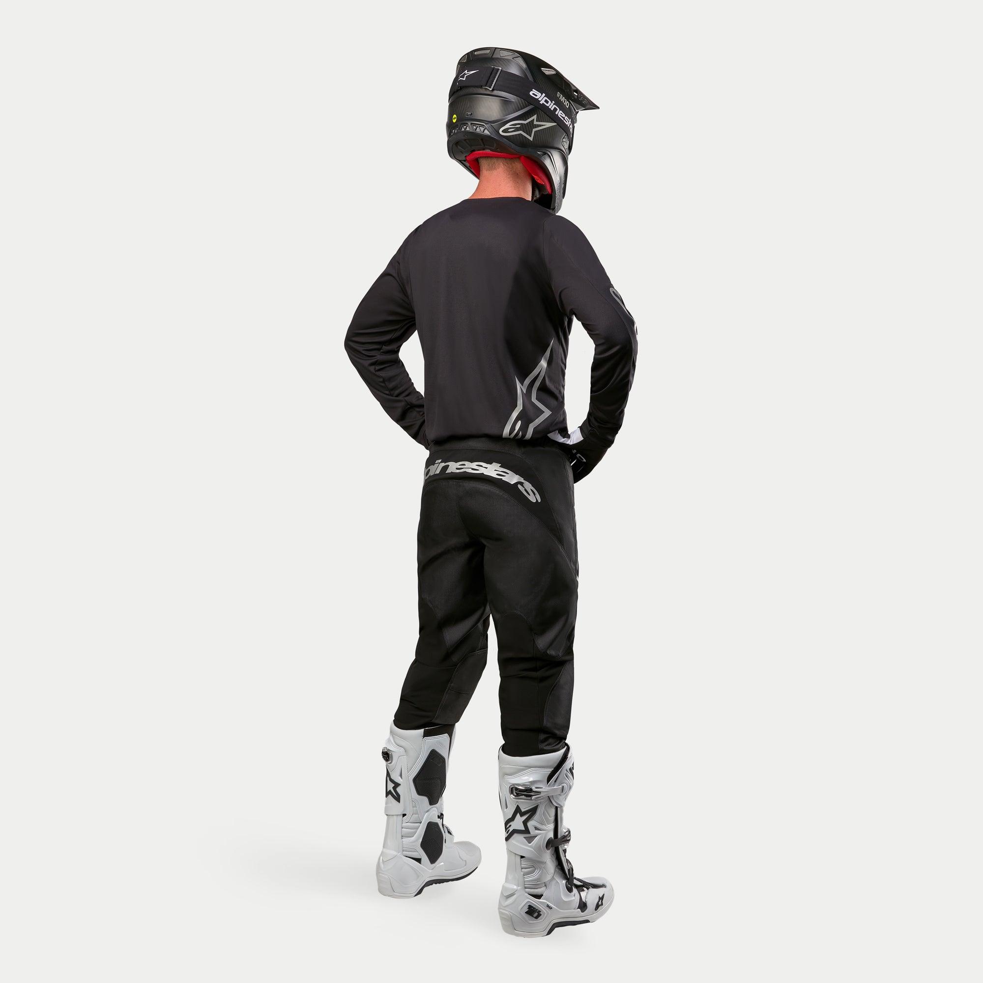 Motocross-Fahrer im Alpinestars 2024 Fluid Graphite Trikot in Schwarz Silber aus der Rückansicht, langärmliges Jersey in athletischer Passform kombiniert mit schwarzer Motocross-Hose, schwarzem Helm und weiß-schwarzen Stiefeln, komplette professionelle Ausrüstung für den Offroad-Rennsport.
