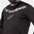 Alpinestars 2024 Fluid Graphite Jersey, Motocross-Jersey, Schwarz und Silber, Nahaufnahme des Brustbereichs mit silbernem Alpinestars-Branding und Stern-Logo, atmungsaktive Poly-Gewebe-Konstruktion, getragen von einem Fahrer mit schwarzem Helm