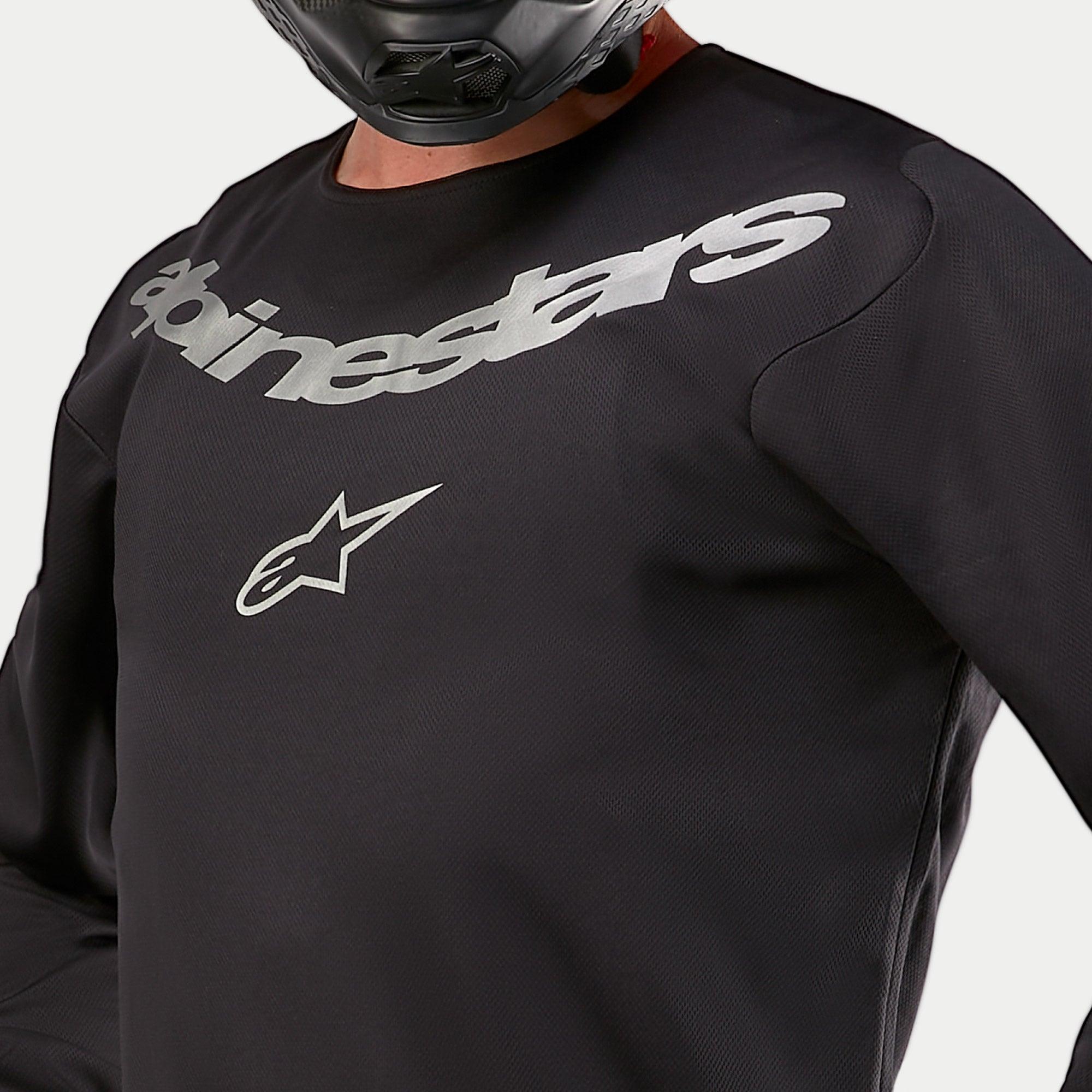 Alpinestars 2024 Fluid Graphite Jersey, Motocross-Jersey, Schwarz und Silber, Nahaufnahme des Brustbereichs mit silbernem Alpinestars-Branding und Stern-Logo, atmungsaktive Poly-Gewebe-Konstruktion, getragen von einem Fahrer mit schwarzem Helm