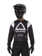 Alpinestars Techdura Trikot, Schwarz, mit einer großen weißen Dreiecks-Brustgrafik und ASTARS-Logo sowie weißen Schulterpartien, getragen von einem Fahrer mit schwarzem Motocross-Helm, Goggles und passender schwarzer Offroad-Ausrüstung.