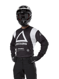 Alpinestars Techdura Jersey, Motocross-Trikot, Schwarz und Weiß, getragen mit passendem Helm und Handschuhen, Pose mit den Händen auf den Hüften vor weißem Hintergrund, mit großem Dreieck-A-Logo und ASTARS-Branding