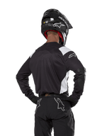 Alpinestars Techdura Trikot, Schwarz mit weißen Schulterakzenten, Rückansicht eines Motocross-Fahrers mit passender schwarzer Hose und Helm, verstärkte Ellbogen-Patches, Enduro- und Offroad-Rennausrüstung